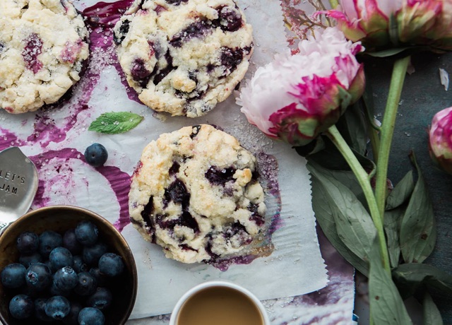 An overhead shot of blueberry cookies beside a bowl of blueberries, flowers, and a cup of coffee 
